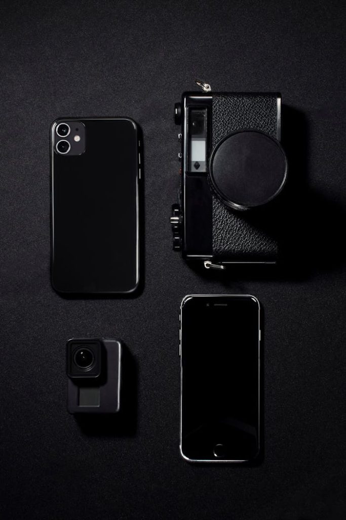 Minimalist flat lay of black gadgets including phones and a camera against a dark background.